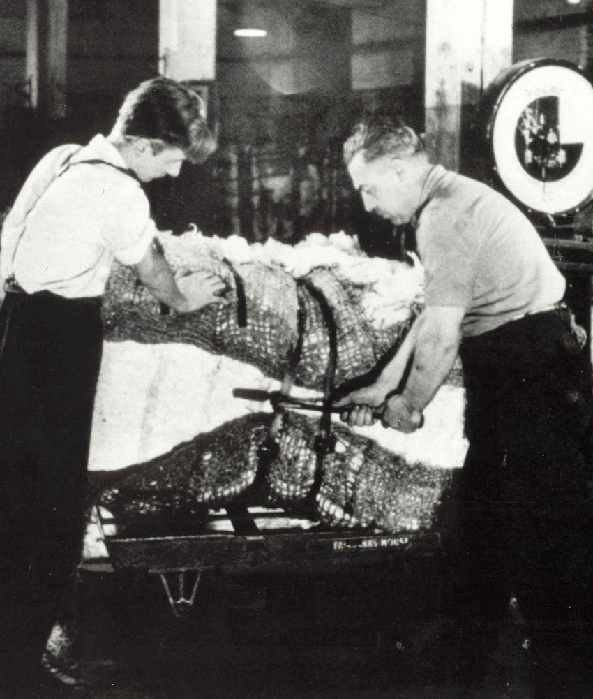 Un ballot de coton au centre, à gauche un homme le tenant et à droite un homme coupe les attaches avec un outil.