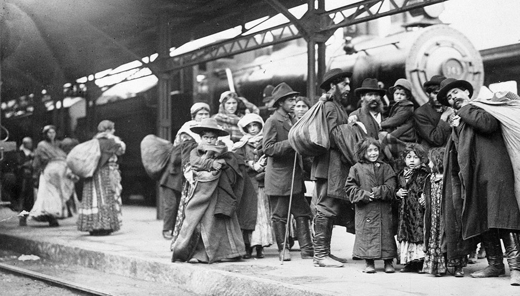 Men, women, and children holding onto their belongings on the platform of a train station. In the background, a locomotive.