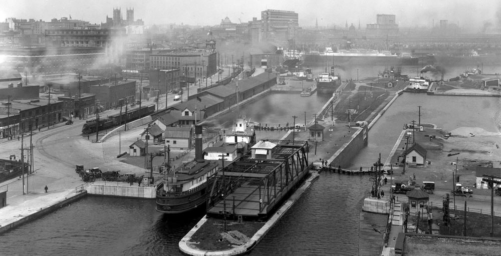 Photo historique du canal Lachine avec bateaux et bâtiments à l'arrière-plan.