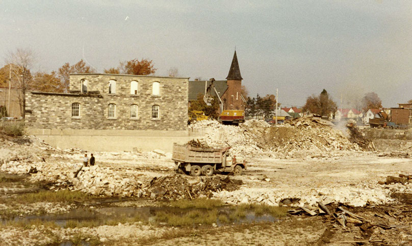 Work site of a building being demolished, mechanical shovel on far side of stone debris; a church in the background.