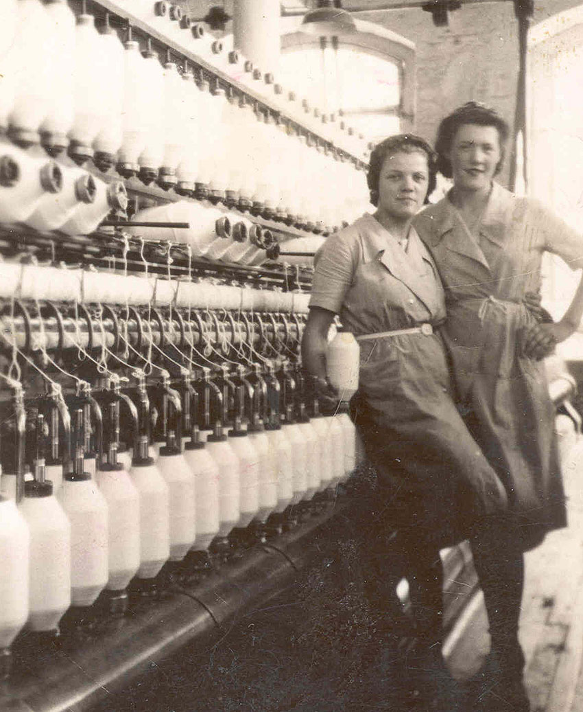 Deux femmes en uniforme devant une machine de filage. Celle de gauche tient une bobine de fil dans sa main droite.