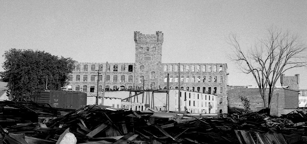 En avant-plan, un empilage de matériaux de démolition, au fond un bâtiment de pierre de cinq étages et une tour.