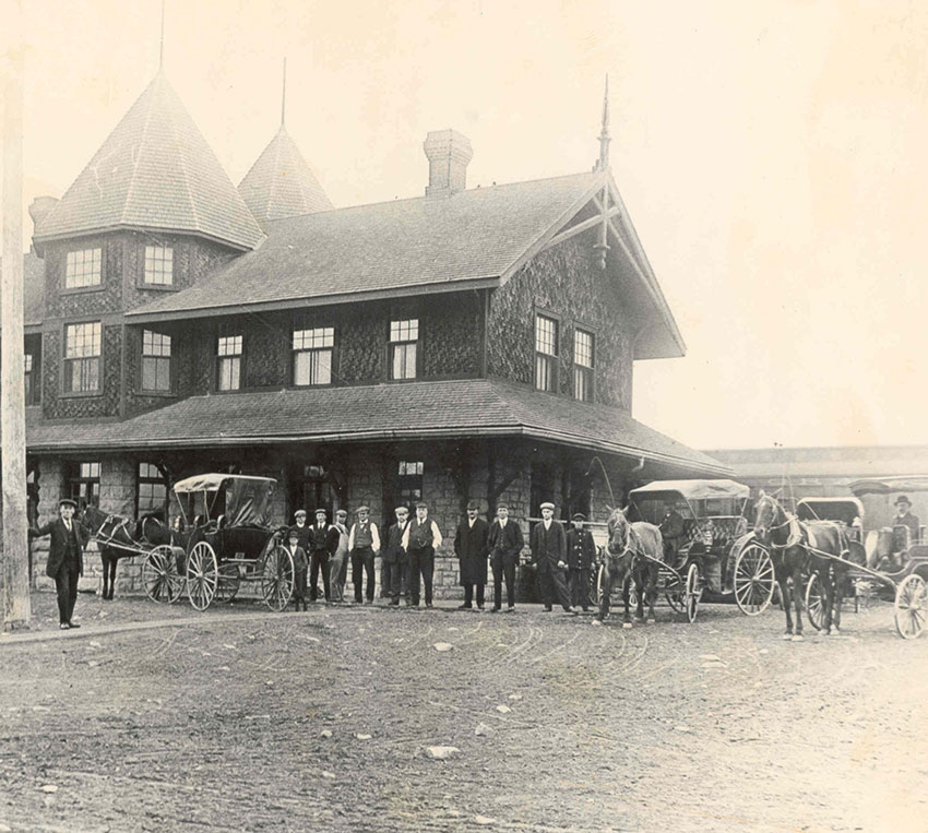 A two-storey railway station with two turrets, surrounded by workers, wagons, and horses.