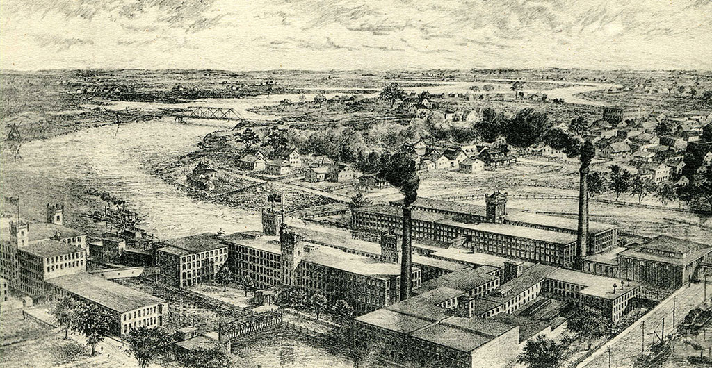 Vue aérienne d’une usine installée sur une rivière avec ses tours, tourelles, cheminées et à gauche un pont de fer.