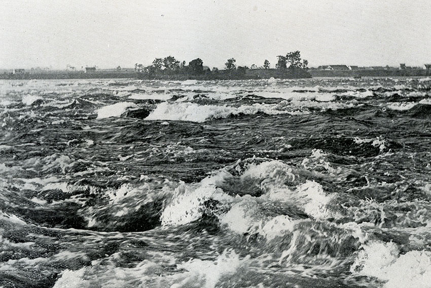 Eddying rapids in a river; in the background, trees and buildings.