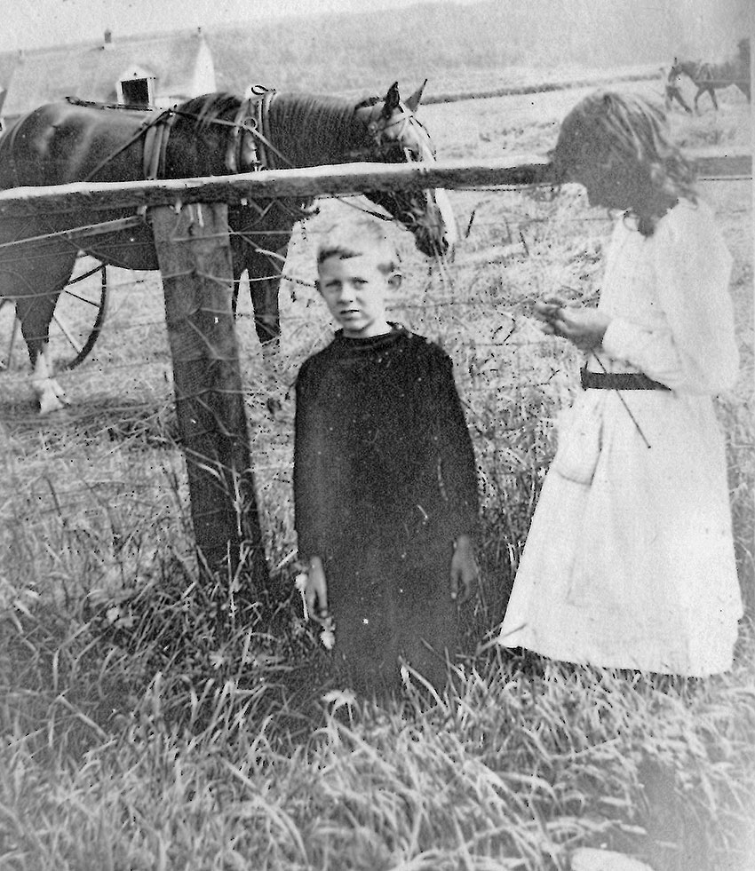 In the foreground, a boy and a girl in front of a fenced-in field; in the background, two horses and a man.