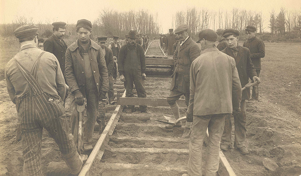 In the centre, workers installing railroad tracks, tools in hand; in the background, trees.
