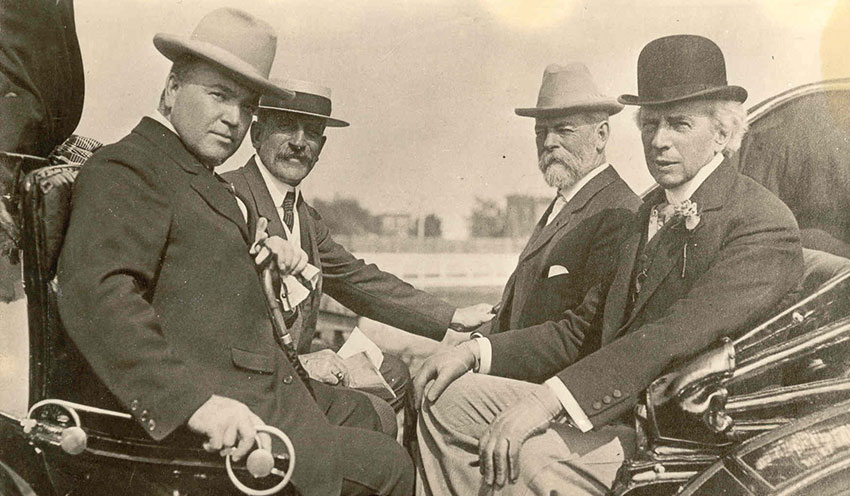 Quatre hommes assis dans une calèche vêtus d’habits et de chapeaux regardant de face.