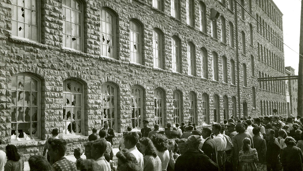A crowd of men, women, and children before a 5- storey building with vandalized windows.