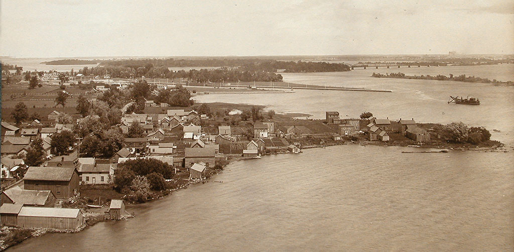 In the foreground, a village on a point of land; in the background, islands, a boat, and a railroad bridge.