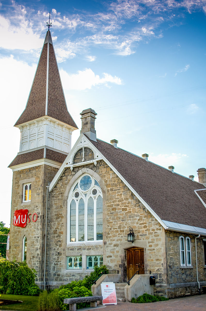 Vieille église en pierre avec un clocher, une verrière et une porte de bois. Sur le clocher, logo MUSO rouge en métal. 